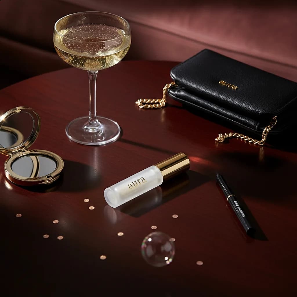 Aura lip gloss on a lounge table with a champagne coupe and a clutch